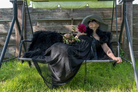 Photo for Portrait of a happy elderly woman 65 - 70 years old in a straw hat resting on a swing in the garden, close up - Royalty Free Image