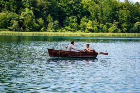 Foto de Plitvice Lakes, Croacia - 30 de agosto de 2021: Pareja joven, un hombre y una niña, navegan en un barco de madera en los lagos de Plitvice y se toman fotos contra el telón de fondo de la naturaleza, Croacia - Imagen libre de derechos