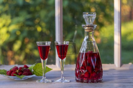 Photo for Homemade red raspberry brandy in two wine glasses and in a glass bottle on a wooden windowsill near summer garden, close up - Royalty Free Image