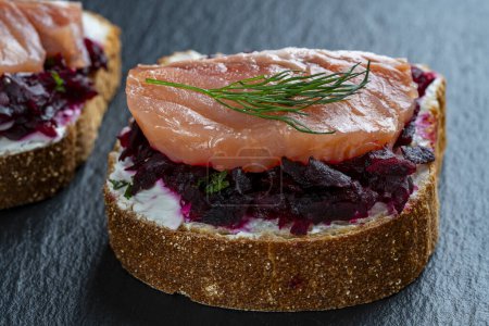 Photo for Toast from a slice of bread with white cream cheese, stewed beetroot pulp,salmon fillets, seeds and green dill on wooden board. Delicious breakfast on morning. Close up - Royalty Free Image