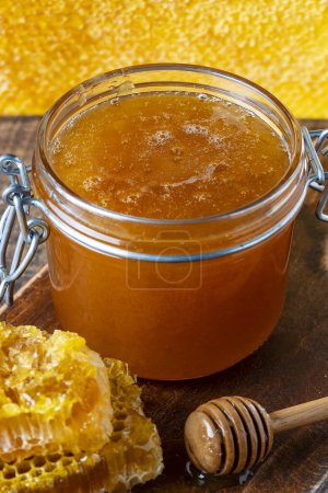 Foto de Un frasco de vidrio de espesa miel dorada con una cuchara de madera y panales. Concepto de apicultura, apicultura, colmenar. Producto dulce de miel, comida saludable. De cerca. - Imagen libre de derechos