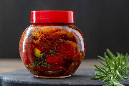 Foto de Tomates rojos secados al sol con ajo, romero verde, aceite de oliva y especias en un frasco de vidrio sobre una mesa de madera. Estilo rústico, de cerca. Tomate seco enlatado - Imagen libre de derechos