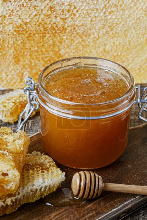Foto de Un frasco de vidrio de espesa miel dorada con una cuchara de madera y panales. Concepto de apicultura, apicultura, colmenar. Producto dulce de miel, comida saludable - Imagen libre de derechos