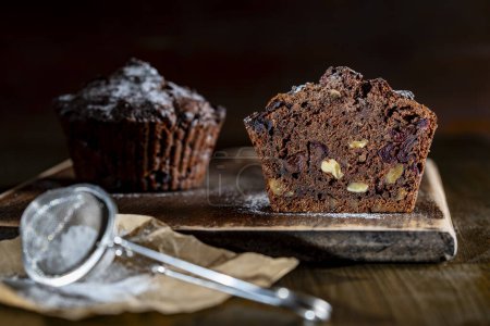 Photo for Chocolate muffin with red cherries and walnuts on a wooden table sprinkled with powdered sugar, close up. Homemade delicious chocolate muffins on wooden board - Royalty Free Image