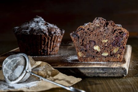 Photo for Chocolate muffin with red cherries and walnuts on a wooden table sprinkled with powdered sugar, close up. Homemade delicious chocolate muffins on wooden board - Royalty Free Image