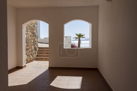 Téléchargez les photos : Trou de fenêtre dans le mur de pierre blanche d'un bâtiment donnant sur la mer Rouge et un palmier sur une plage tropicale dans la station balnéaire de Sharm El Sheikh, Egypte - en image libre de droit