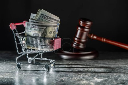 Photo for Shopping cart on wheels with dollar bills and wooden judge gavel on dark background, close up. Finance, business, purchases, spending and sales concept - Royalty Free Image
