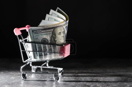Photo for Shopping cart on wheels with dollar bills in it on dark background, close up, copy space. Finance, business, purchases, spending and sales concept - Royalty Free Image