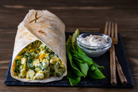 Foto de Burrito casero envuelve con huevos cocidos, ajo verde silvestre y crema agria para el desayuno saludable sobre tabla de madera, de cerca - Imagen libre de derechos