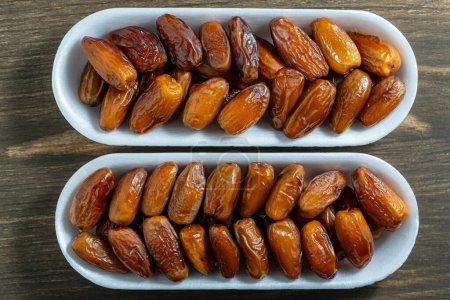 Foto de Frutas dátiles secas en un recipiente blanco de dos sobre fondo de madera, vista superior, primer plano - Imagen libre de derechos