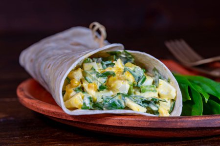 Foto de Burrito casero envuelve con huevos cocidos, patatas, ajo verde silvestre y crema agria para el desayuno saludable en el plato, de cerca - Imagen libre de derechos