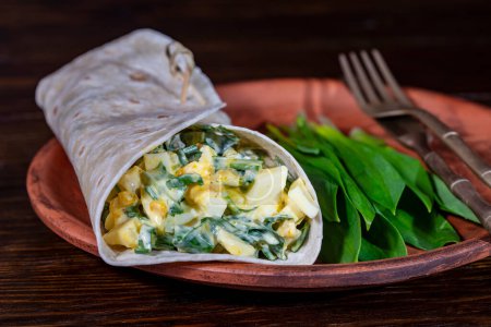 Foto de Burrito casero envuelve con huevos cocidos, patatas, ajo verde silvestre y crema agria para el desayuno saludable en el plato, de cerca - Imagen libre de derechos
