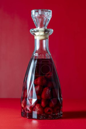 Glasflasche hausgemachten roten Himbeerlikör auf rotem Hintergrund, Nahaufnahme. Konzept für alkoholische Getränke