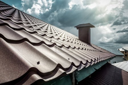Brown corrugated metal profile roof installed on a modern house. The roof of corrugated sheet. Roofing of metal profile wavy shape under cloudy sky and sunlight