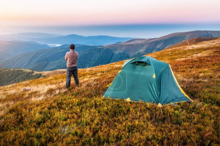 Photo for Tourist hiker standing near tent and looking on mountains at sunset. Travel and vacation theme - Royalty Free Image