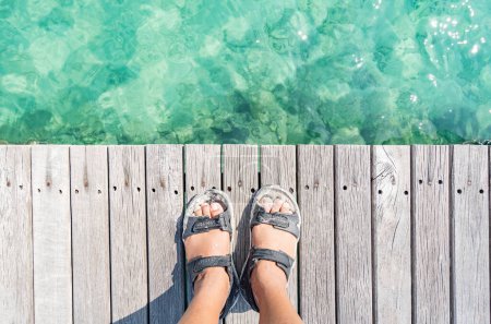 Foto de Los pies de un hombre de pie sobre una tabla de madera junto al mar, agua de mar verde brillante, Koh Mak, provincia de Trat, Tailandia. - Imagen libre de derechos