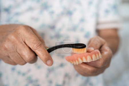 Photo for Asian elderly woman patient use toothbrush to clean partial denture of replacement teeth. - Royalty Free Image