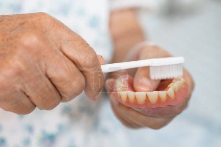 Téléchargez les photos : Asiatique femme âgée patient utiliser une brosse à dents pour nettoyer une prothèse partielle de dents de remplacement. - en image libre de droit