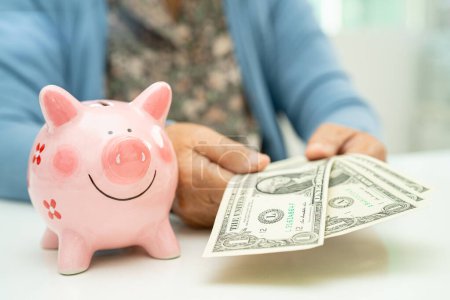 Asiática anciana sosteniendo billetes en dólares con alcancía rosa para ahorrar dinero y seguros, pobreza, problemas financieros en la jubilación.