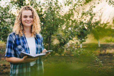 Photo for Smiling woman with curly-hair using modern technologies in agriculture orchard writing notes in her notebook. Free space for text. Man agronomist farmer with - Royalty Free Image