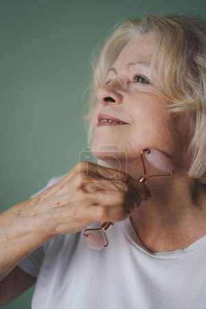 Téléchargez les photos : Portrait en gros plan d'une femme âgée faisant un massage facial avec un rouleau de jade, fermant les yeux. Jolie femme propre peau fraîche. Traitement antiâge. Beauté femme - en image libre de droit
