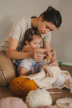 Foto de Hermosa madre enseñando a su hija cómo tejer patrones para la temporada de otoño en la sala de estar en casa. Participar en la actividad de hobby en fin de semana - Imagen libre de derechos