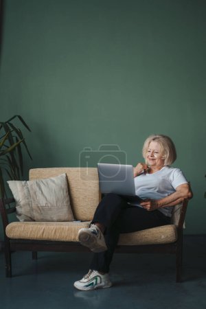 Foto de Mujer mayor con parches debajo de los ojos sentada en el sofá frente a la computadora portátil durante el trabajo remoto. Retrato contemporáneo. Generación de ancianos en línea. Retrato de belleza - Imagen libre de derechos