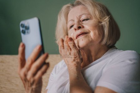 Foto de Mujer mayor alegre con parches de colágeno bajo los ojos sentada con su teléfono inteligente en el sofá por la mañana. Retrato contemporáneo. - Imagen libre de derechos
