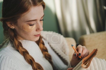 Foto de Mujer joven y atractiva con ojos haciendo rutina matutina de cuidado facial mientras toma notas en su diario. Mantener un diario - Imagen libre de derechos