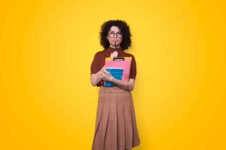 Portrait of thinking female student holding pencil and notebooks, looking at camera, isolated over yellow background. Positive person. Poster 625513168