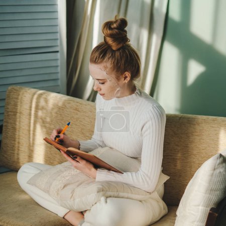 Foto de Mujer pelirroja haciendo tratamiento de belleza facial escribiendo su diario mientras se relaja sentada en su cómodo sofá. Rutina de cuidado de la piel. Cuidado facial de la piel. Mujer. - Imagen libre de derechos