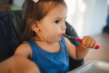 Retrato caucásico bebé niña comiendo pasta de plato sentado trona, comida sólida para bebés. Concepto de autoalimentación