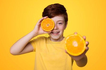 Téléchargez les photos : Garçon tenant deux tranches d'orange dans ses mains, il couvre son oeil avec l'un et tient l'autre vers la caméra isolée sur fond jaune. - en image libre de droit
