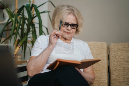 Photo for Beautiful senior woman writing information in notebook while sitting on sofa at home. Retirement hobby for training memory. Alzheimers preventive practice. - Royalty Free Image