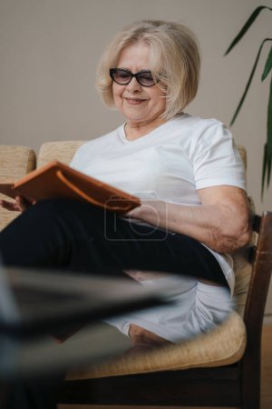Photo for Elderly minded wom sitting on sofa writing down in notebook staying at home, spend free spare time in living room. Happy lifestyle. Apartment interior. - Royalty Free Image