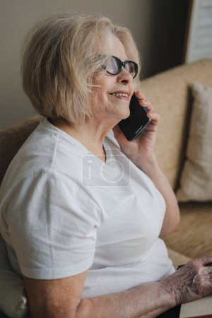 Photo for Senior woman talking with client on smartphone and writing in notepad while sitting on sofa at home. Having fun at home, sitting on couch - Royalty Free Image