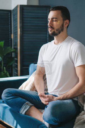 Foto de Hombre guapo practicando meditación en la postura de piernas cruzadas, en el sofá de su sala de estar. Ejercicio respiratorio. - Imagen libre de derechos