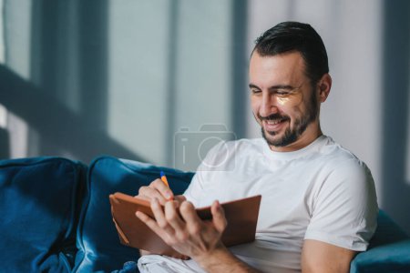 Photo for Portrait of a positive young businessman sitting on the sofa writing in notebook relaxing and resting at home. Conept of wellness, daily routine and self - Royalty Free Image