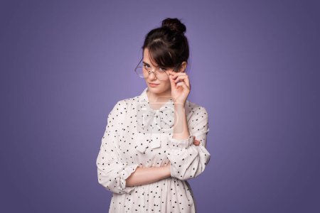 Photo for Studio portrait of serious focused woman with eyeglasses, attentively looking over her glasses, touching eyeglasses frames with hand, standing over purple - Royalty Free Image