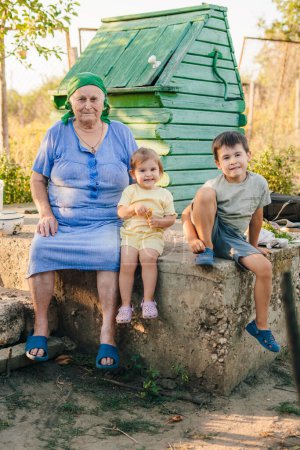 Foto de Happy grandmother sitting near baby gir and boy outdoors in the garden. Garden, family and grandparent. Care and happiness together. - Imagen libre de derechos