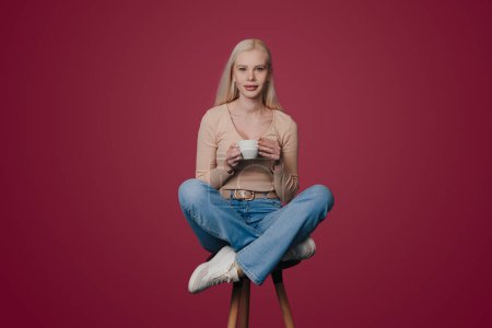 Photo for Full length body size photo of woman holding cup of cappuccino sitting on chair and looking at camera isolated on crimson color background. - Royalty Free Image