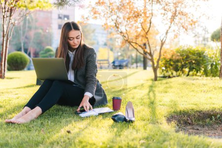 Foto de Mujer freelancer en ropa casual sentada en el césped en el parque escribiendo sus notas en su cuaderno con su computadora en sus piernas. Trabajo independiente con - Imagen libre de derechos