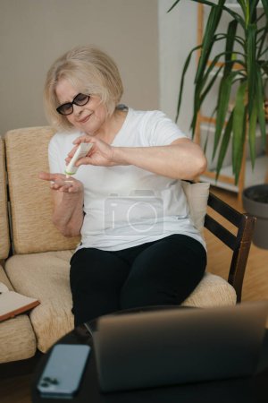 Téléchargez les photos : Portrait d'une femme âgée hydratant sa peau des mains assise sur un canapé à la maison. Peau de beauté. - en image libre de droit