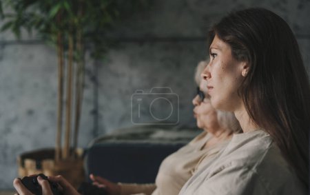 Photo for Side view of a woman with her mother playing games on TV, sitting on cozy sofa. Healthy activity. Happy cheerful family. Happy lifestyle. Family home leisure. - Royalty Free Image