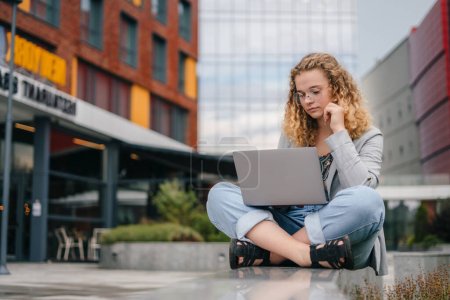 Photo for Photo of beautiful curly haired woman sitting outdoors in front of university campus holding notebook typing essay text wearing eyeglasses. Online communication - Royalty Free Image