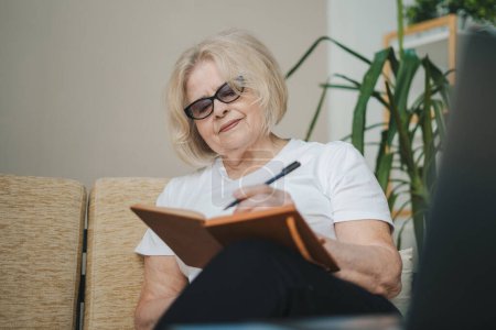 Photo for Elderly woman wears casual clothes sitting on sofa writing down in notebook diary, staying at home, spend free spare time in living room. Modern old senior - Royalty Free Image