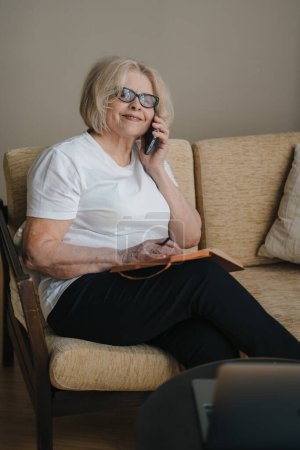 Foto de Mujer mayor sonriente sentada en el sofá leyendo información en su planificador o diario y haciendo una llamada telefónica. - Imagen libre de derechos