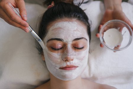 Draufsicht der Ernte anonyme weibliche Anwendung Gesichtsmaske auf Gesicht mit Pinsel, während im hellen Raum sitzen