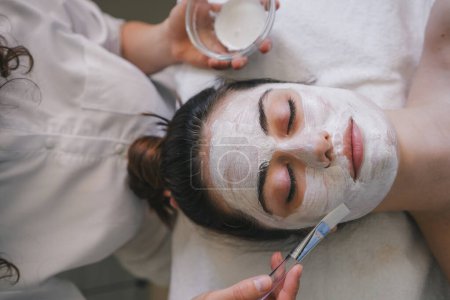 Téléchargez les photos : Femme caucasienne faisant un traitement de beauté au salon de spa et étant appliqué crème sur son visage. Masque peeling visage, soin de beauté spa, soins de la peau. - en image libre de droit