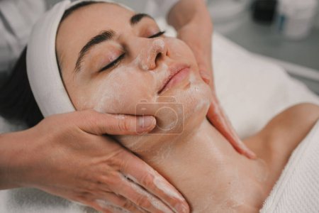 Foto de Mujer bonita joven que recibe tratamientos usando espuma de limpieza facial en salones de belleza. Tratamiento de masaje corporal. Tratamiento facial, cosmetología. - Imagen libre de derechos
